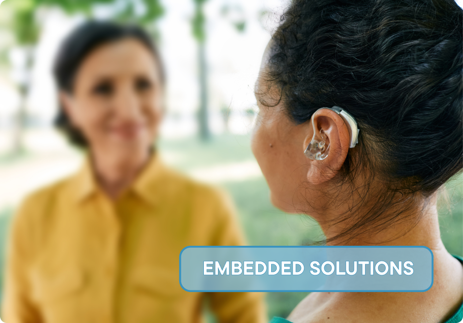 A picture from behind of a young woman, who's face is not visible. She is wearing a hearing aid and speaking to an older woman. Overlayed with the sticker "Embedded Solutions".