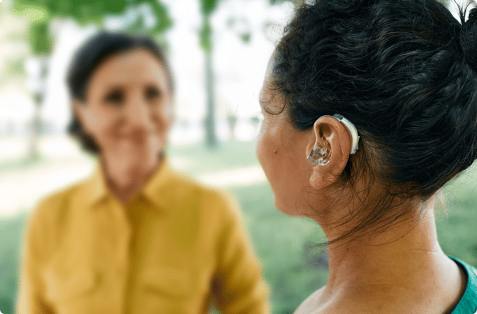 Picture of a young woman wearing a hearing aid, while talking to an older woman in blurred view. 